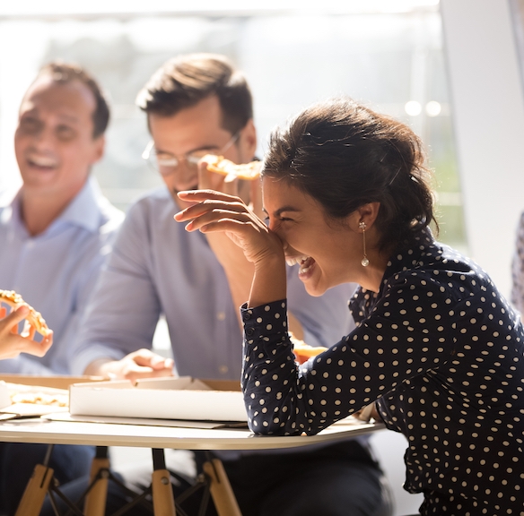 Indian woman laughing at funny joke eating pizza with diverse coworkers in office